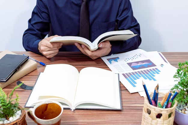 A person reading a book on a table surrounded by documents and data